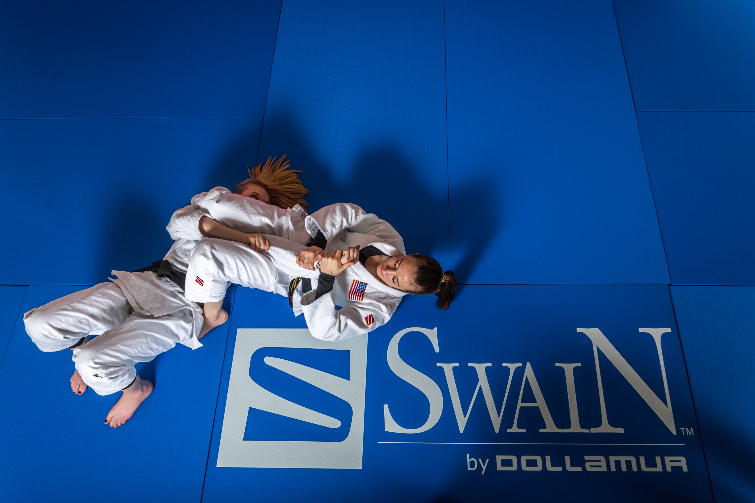 Two people in white martial arts uniforms grappling on a blue jiu jitsu mat mat with Swain by Dollamur branding.